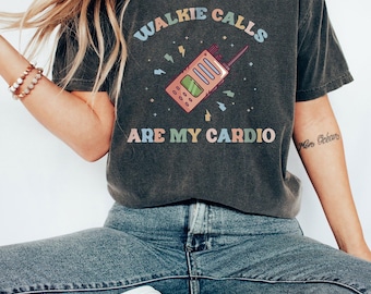 May include: A charcoal gray short-sleeve t-shirt featuring a graphic of a pink walkie-talkie with the text "WALKIE CALLS ARE MY CARDIO" in a retro, multi-colored font, surrounded by small lightning bolt and star icons.