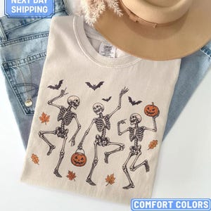 May include: An ivory t-shirt featuring a graphic of three dancing skeletons, orange jack-o'-lanterns, and scattered autumn leaves. The design includes black bats flying above the skeletons. The shirt is paired with blue denim jeans and a tan hat.