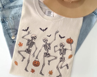 Può includere: Una t-shirt color avorio con una grafica di tre scheletri danzanti, zucche di Halloween arancioni e foglie autunnali sparse. Il design include pipistrelli neri che volano sopra gli scheletri. La t-shirt è abbinata a jeans blu e un cappello beige.