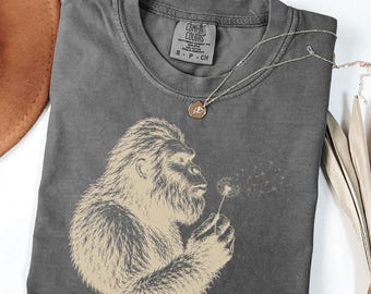 Può includere: Una t-shirt girocollo grigio antracite con una stampa beige chiaro raffigurante una creatura pelosa che soffia su un dente di leone. La maglietta presenta l'etichetta del marchio Comfort Colors all'interno del collo ed è abbinata a una catenina.