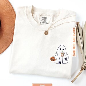 May include: An ivory t-shirt featuring a graphic of a white ghost holding an orange jack-o'-lantern and a plastic cup with a straw. The shirt has a Comfort Colors brand label at the neck, and a gold-toned pendant necklace rests on the fabric.