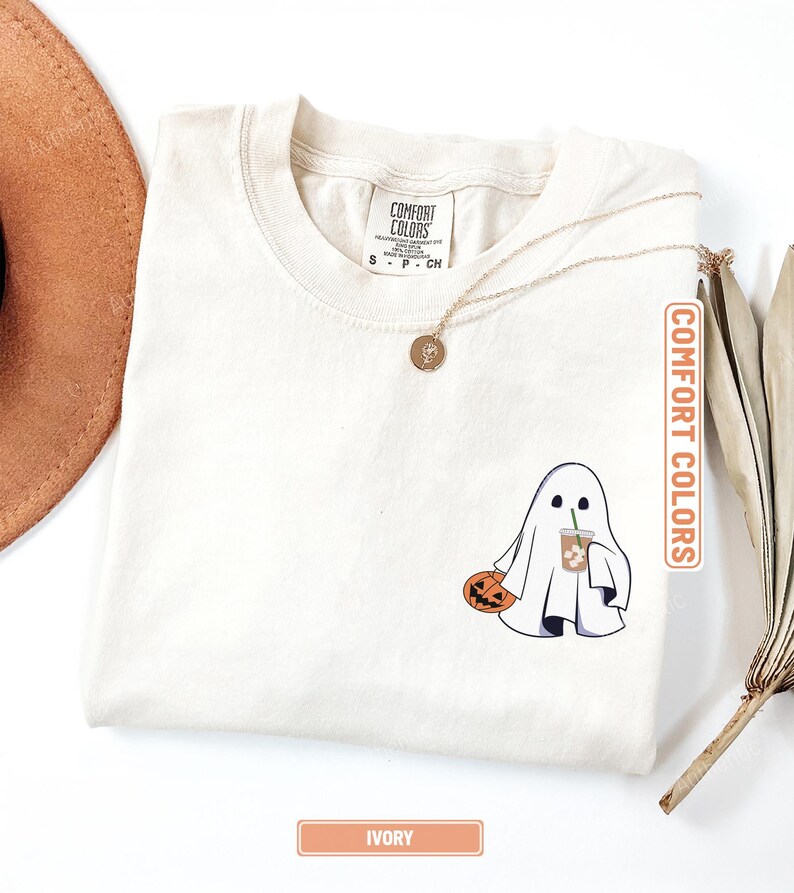 May include: An ivory t-shirt featuring a graphic of a white ghost holding an orange jack-o'-lantern and a plastic cup with a straw. The shirt has a Comfort Colors brand label at the neck, and a gold-toned pendant necklace rests on the fabric.