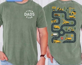 Può includere: Una t-shirt a maniche corte color verde salvia con un logo circolare bianco sul petto che recita TIRED DADS CLUB. Il retro presenta una grafica di una strada nera tortuosa con veicoli da cantiere gialli, edifici e alberi.