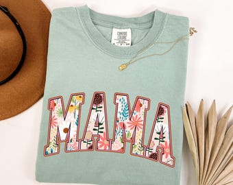 Puede incluir: Una camiseta de color verde salvia con la palabra MAMA en letras grandes. Las letras están rellenas con un colorido estampado floral de flores y hojas rosas, blancas, amarillas y verdes. La camiseta tiene una etiqueta de la marca Comfort Colors.
