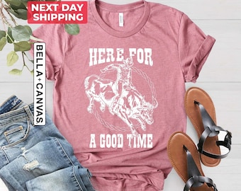May include: A heather mauve t-shirt featuring a white graphic of a cowboy riding a bucking bull inside a rope circle. The text "HERE FOR A GOOD TIME" is printed in white above and below the graphic. The shirt has short sleeves and a crew neck.