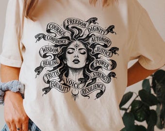 Può includere: Una t-shirt color avorio con una stampa grafica nera di una donna con serpenti al posto dei capelli. Il design include le parole Freedom, Dignity, Autonomy, Justice, Power, Strength, Resilience, Voice, Equality, Truth, Healing, Wisdom, Confidence e Courage.