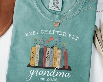 Può includere: Una t-shirt verde chiaro con la grafica di una fila di libri su uno scaffale con fiori che crescono dalla parte superiore. Il testo recita BEST CHAPTER YET, grandma e est 2026. I dorsi dei libri riportano frasi come ENDLESS HUGS e FULL HEARTS.