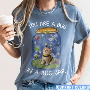 May include: A blue t-shirt featuring a graphic of a kitten wearing a bee costume inside a glass jar. The jar contains illustrations of a ladybug, dragonfly, butterfly, grasshopper, and bee. The text reads, "YOU ARE A BUG IN A BUG JAR."
