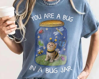 Può includere: Una t-shirt blu con la grafica di un gattino vestito da ape all'interno di un barattolo di vetro. Il barattolo contiene illustrazioni di coccinella, libellula, farfalla, cavalletta e ape. Il testo recita: "YOU ARE A BUG IN A BUG JAR."