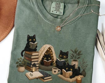 Può includere: Una t-shirt verde muschio con una grafica di cinque gatti neri che leggono libri in un ambiente accogliente con una piccola tenda, piante in vaso, una lanterna e una tazza. La maglietta presenta l'etichetta Comfort Colors sul colletto.