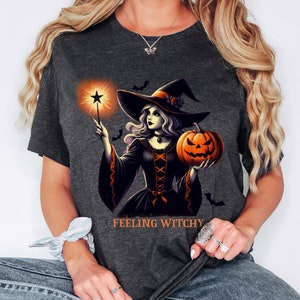 May include: Dark gray t-shirt featuring a Halloween witch illustration. The witch wears a black dress, a pointed hat, and holds a glowing star wand and a carved pumpkin. The text "FEELING WITCHY" is printed below the illustration.
