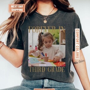 May include: A charcoal grey t-shirt featuring a square photographic print of a child drawing at a desk with coloured pencils. The text "FOREVER IN" appears above the photo, and "THIRD GRADE" is printed below in a gold serif font.