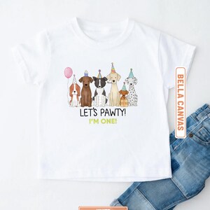 May include: White Bella Canvas t-shirt featuring a colorful illustration of seven dogs wearing party hats and holding a balloon. The text "LET'S PAWTY! I'M ONE!" is printed below the dogs. Blue jeans are visible.