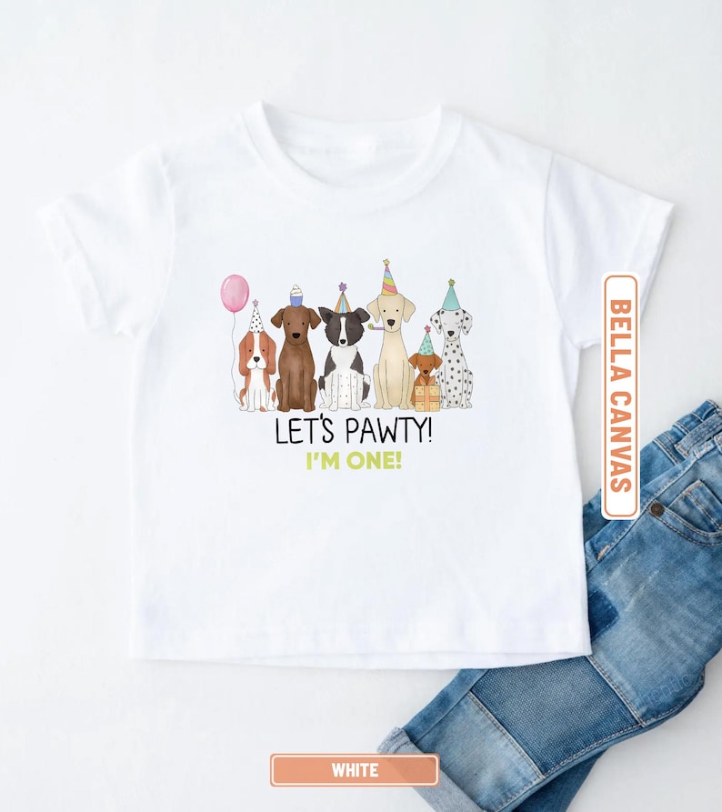 May include: White Bella Canvas t-shirt featuring a colorful illustration of seven dogs wearing party hats and holding a balloon. The text "LET'S PAWTY! I'M ONE!" is printed below the dogs. Blue jeans are visible.