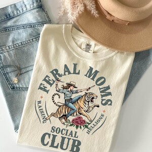 May include: A cream-colored t-shirt featuring a graphic of a person riding a tiger while holding knives. The text reads FERAL MOMS RAISING HELLIONS SOCIAL CLUB. The design includes floral accents and a tan hat and blue denim jeans are visible.
