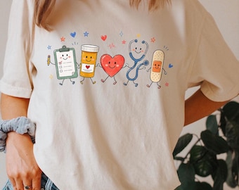 Può includere: Una t-shirt color avorio con una stampa grafica di cinque oggetti medici antropomorfi: una cartellina, un flacone di pillole, un cuore rosso, uno stetoscopio blu e un cerotto beige, tutti con facce sorridenti e piccole stelle colorate.