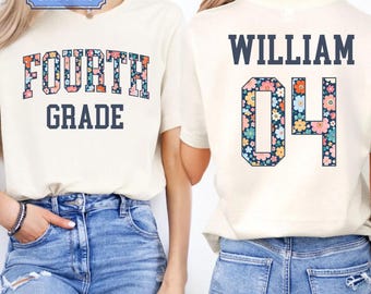 Può includere: Una t-shirt color naturale con il testo FOURTH GRADE in un carattere universitario blu scuro con riempimento a motivo floreale sul davanti, e il nome WILLIAM con il numero 04 in un corrispondente stile universitario a motivi floreali sul retro.