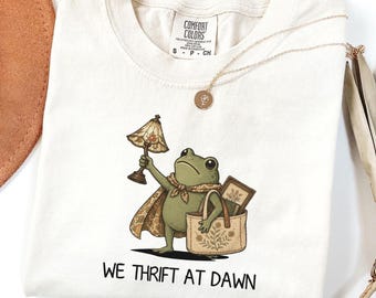 Può includere: Una t-shirt color avorio con la grafica di una rana verde che indossa un mantello e una sciarpa fantasia, tiene in mano una lampada vintage e una borsa con motivo floreale. Il testo sotto la rana recita WE THRIFT AT DAWN. Etichetta Comfort Colors.