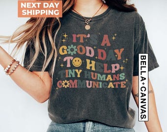 May include: A charcoal gray short-sleeve t-shirt featuring the multi-colored text IT'S A GOOD DAY TO HELP TINY HUMANS COMMUNICATE, accented with a yellow smiley face, small flower icons, and star shapes printed on the front of the garment.