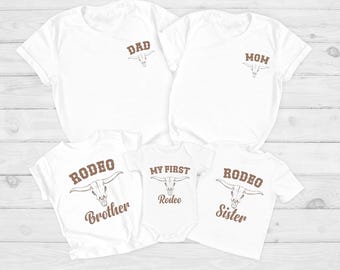 May include: A set of five white t-shirts and a baby bodysuit featuring brown text and a bull skull graphic. The shirts are labeled DAD, MOM, RODEO Brother, and RODEO Sister, while the baby bodysuit is printed with MY FIRST Rodeo.