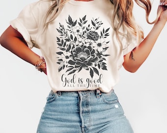 May include: A cream-colored short-sleeve t-shirt featuring a black graphic of a floral bouquet with leaves and blossoms. Below the floral design, the text reads "God is good" in a script font, with "ALL THE TIME" printed in smaller capital letters.
