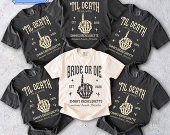 Può includere: Cinque magliette a maniche corte in nero e crema. Quattro magliette nere presentano una grafica con mano scheletrica e il testo 'TIL DEATH do us party', 'EST 2026' e 'EMMA'S BACHELORETTE miami beach, florida'. Una maglietta crema dice 'BRIDE OR DIE'.