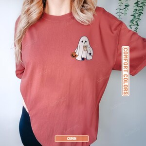 May include: A cumin-colored short-sleeve t-shirt featuring a graphic of a white ghost holding an orange pumpkin and a brown iced coffee. The shirt includes the text COMFORT COLORS and CUMIN printed on rectangular labels on the right side.