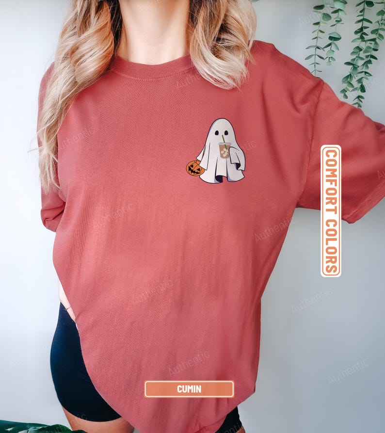 May include: A cumin-colored short-sleeve t-shirt featuring a graphic of a white ghost holding an orange pumpkin and a brown iced coffee. The shirt includes the text COMFORT COLORS and CUMIN printed on rectangular labels on the right side.
