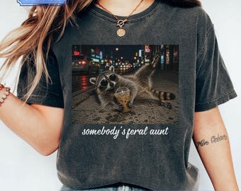 Può includere: Una t-shirt grigio antracite con la grafica di un procione che indossa occhiali da sole rotondi e beve da un bicchiere di plastica con una cannuccia. Sotto l'immagine, il testo recita somebody's feral aunt in un carattere corsivo bianco.