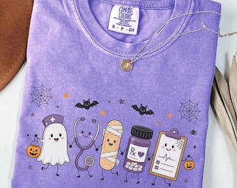 Può includere: Una t-shirt viola con una grafica raffigurante un fantasma, uno stetoscopio, una benda, un flacone di medicinale con etichetta Rx e una cartellina. Il design include piccoli pipistrelli, ragnatele e zucche. La maglia ha un'etichetta Comfort Colors.