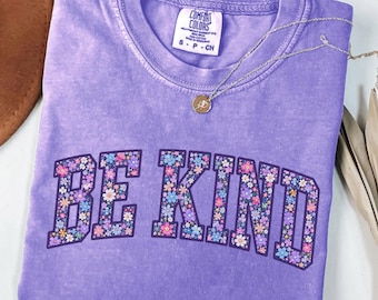 Può includere: Una t-shirt viola a maniche corte con scollo rotondo. La parte anteriore presenta la scritta BE KIND in grandi lettere stampatello riempite con un colorato motivo floreale di piccoli fiori rosa, blu e bianchi. Una collana argentata.