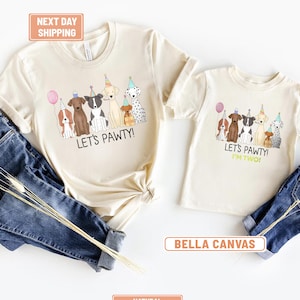 May include: Two natural-colored t-shirts with a dog-themed graphic. The larger shirt says "LET'S PAWTY!" and the smaller shirt says "LET'S PAWTY! I'M TWO!". The graphic features cartoon dogs wearing party hats and holding a balloon.