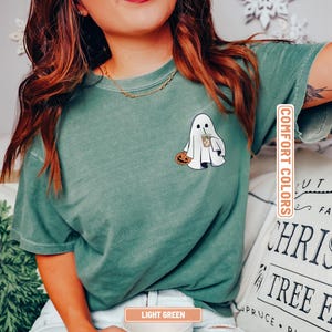 May include: A sage green short-sleeve t-shirt featuring a graphic on the left chest of a white ghost holding an orange jack-o'-lantern and a brown iced coffee drink. The shirt has a crew neckline and a vintage-style washed fabric texture.