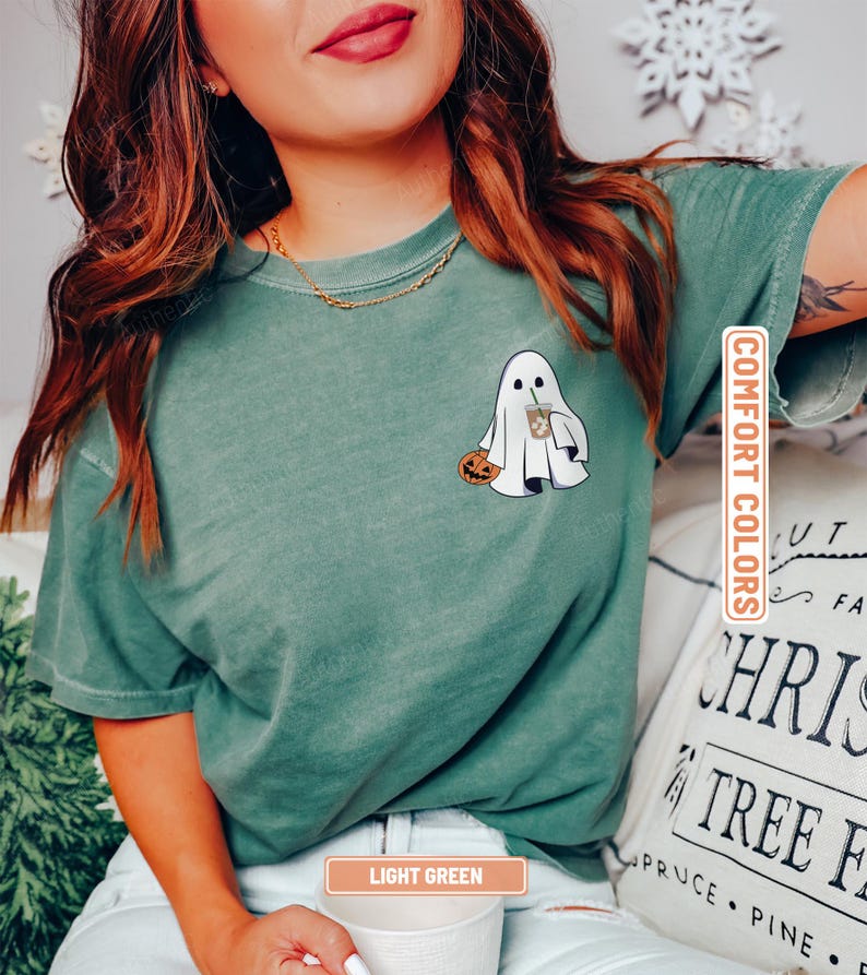 May include: A sage green short-sleeve t-shirt featuring a graphic on the left chest of a white ghost holding an orange jack-o'-lantern and a brown iced coffee drink. The shirt has a crew neckline and a vintage-style washed fabric texture.