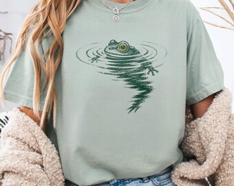 Può includere: Una t-shirt a maniche corte color verde salvia con una stampa grafica verde scuro di una rana parzialmente immersa nell'acqua, che crea increspature sulla superficie. La rana ha un occhio giallo prominente. Girocollo e vestibilità comoda.
