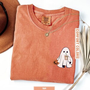 May include: A yam-colored short-sleeve t-shirt with a crew neckline. The shirt features a graphic of a white ghost holding a jack-o'-lantern and an iced coffee. The inner neck label reads Comfort Colors. A gold chain necklace rests on the fabric.