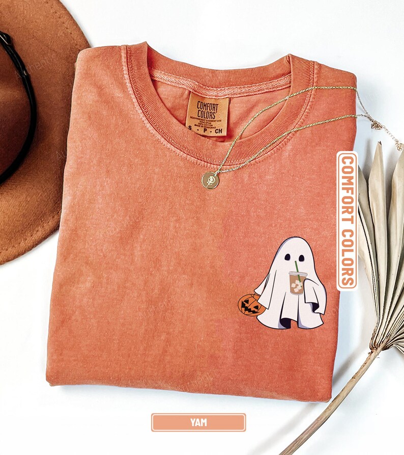 May include: A yam-colored short-sleeve t-shirt with a crew neckline. The shirt features a graphic of a white ghost holding a jack-o'-lantern and an iced coffee. The inner neck label reads Comfort Colors. A gold chain necklace rests on the fabric.
