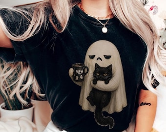 Può includere: Una t-shirt nera con la grafica di un fantasma beige che tiene in mano una tazza nera con la scritta MEH e una piccola icona di fantasma, mentre tiene in braccio un soffice gatto nero. Il fantasma ha un'espressione imbronciata e il gatto occhi gialli.