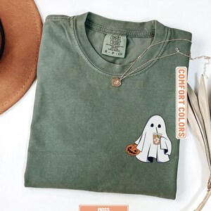 May include: A moss green short-sleeve t-shirt with a crew neckline. The shirt features a graphic of a white ghost holding a jack-o'-lantern and an iced coffee. The interior neck label reads Comfort Colors and the size S-P-CH. A thin gold chain necklace rests on the fabric.