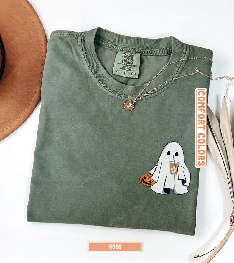 May include: A moss green short-sleeve t-shirt with a crew neckline. The shirt features a graphic of a white ghost holding a jack-o'-lantern and an iced coffee. The interior neck label reads Comfort Colors and the size S-P-CH. A thin gold chain necklace rests on the fabric.