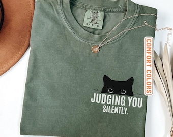 May include: A moss green short-sleeve t-shirt featuring a black cat graphic peeking over the text "JUDGING YOU SILENTLY." The shirt has a crew neckline and a Comfort Colors brand label on the inside collar. A thin gold-tone necklace rests on the fabric.