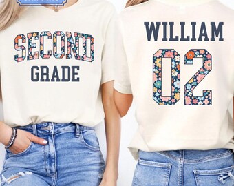 Può includere: Una t-shirt color naturale con la scritta SECOND GRADE sul davanti in lettere stampatello blu scuro con riempimento a motivo floreale. Il retro mostra il nome WILLIAM sopra il numero 02, anch'esso in blu scuro con un motivo floreale.