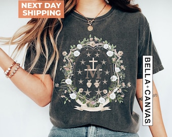 May include: A charcoal gray short-sleeve t-shirt featuring a central graphic of a floral wreath surrounding a cross, the letter M, and two hearts. The design includes small stars and a ribbon at the bottom. The shirt has a crew neckline.