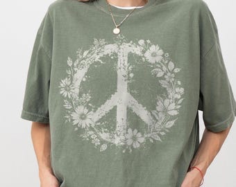 Può includere: Una t-shirt a maniche corte color verde muschio con una grande grafica bianca dall'aspetto vissuto raffigurante un simbolo di pace circondato da fiori e foglie. Il disegno è centrato sul petto e la maglia ha una vestibilità ampia.