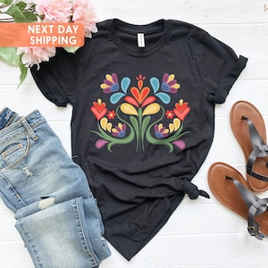 May include: Black t-shirt featuring a colourful floral design. The design incorporates a symmetrical pattern of flowers and leaves in shades of red, yellow, blue, green, and purple.