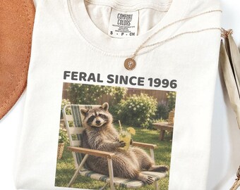 Può includere: Una t-shirt color avorio con la grafica di un procione seduto su una sedia da giardino che tiene in mano un bicchiere di limonata. Il testo sopra la grafica recita FERAL SINCE 1996. La maglietta ha un'etichetta Comfort Colors sul colletto.