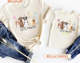 Può includere: Due magliette color naturale con una grafica di sei cani che indossano cappellini da festa, con il testo "LET'S PAWTY!" su entrambe e "I'M TWO!" sulla più piccola. Le magliette sono abbinate a dei jeans blu.