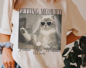 Può includere: Una t-shirt color avorio con una grafica in bianco e nero di un gatto che indossa occhiali da sole a forma di cuore e un velo da sposa in pizzo. Il testo recita GETTING MEOWIED, Future Mrs. William e EST. 2026. Il gatto ha una zampa alzata.
