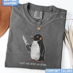 May include: A dark grey short-sleeve t-shirt featuring a graphic of a penguin holding a knife. Below the penguin, the text reads "FLIGHT WAS NEVER AN OPTION". The shirt has a crew neckline and a tag printed on the inside collar area.