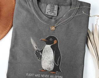 Può includere: Una t-shirt a maniche corte grigio scuro con la grafica di un pinguino che tiene in mano un coltello. Sotto il pinguino, il testo recita "FLIGHT WAS NEVER AN OPTION". La maglietta ha un girocollo e un'etichetta stampata all'interno del colletto.